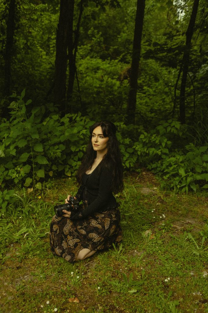 Hailey, owner of Hailey Woods Photography sitting in the grass posing with her camera. 
