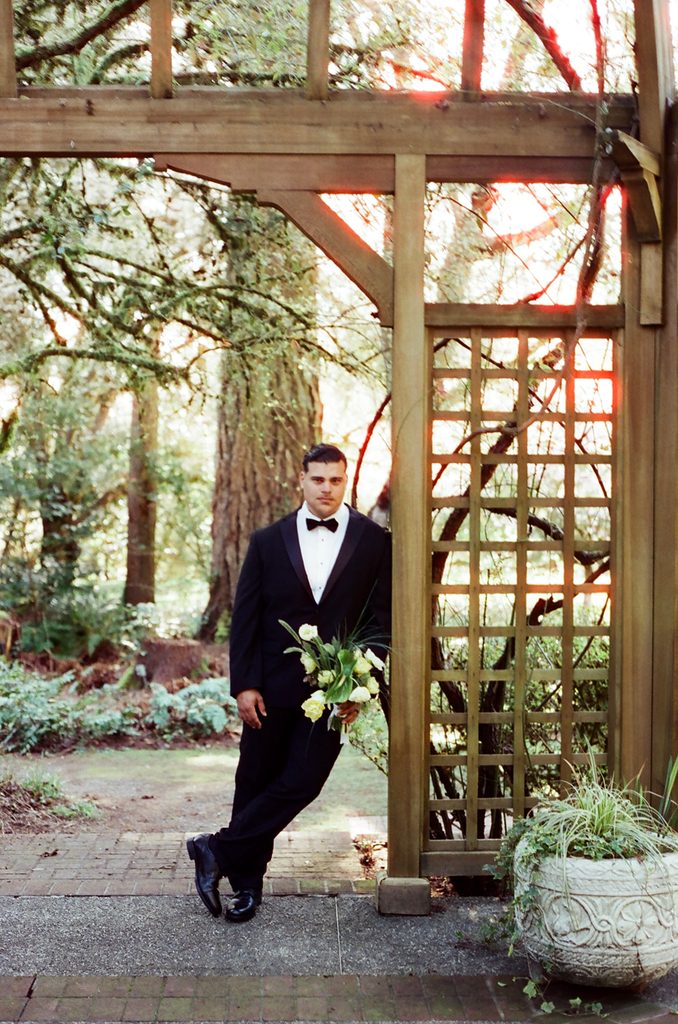 Groom in a black tuxedo holding a bouquet beneath a wooden garden structure at Lakewold Gardens, photographed on CineStill 400D film.